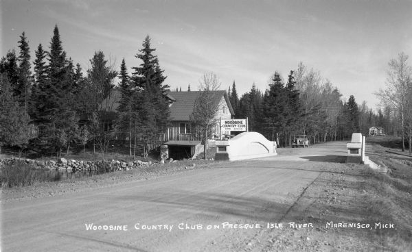 View of the Woodbine Country Club on Presque Isle River. An automobile is parked on the side of the road near the County Club sign which says “Woodbine Country Club, Dine, Dance.” There is a dirt road, river, and bridge in the foreground.