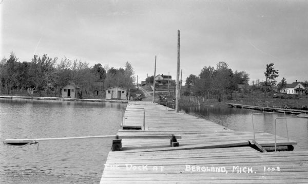 View down long wooden dock. There is a diving board on the dock as well as ladders for people swimming. On shore are three parked automobiles and a few cabins or storage buildings. Also visible are trees, utility poles, dirt roads and other buildings.