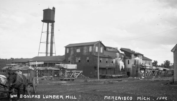 View of William Bonifas Lumber Mill showing the mill buildings, a water tower, "Railroad Crossing" sign, an automobile, lumber stacked on railroad cars, and a man driving a team of horses with a wagon full of lumber.
