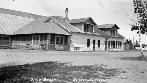 Exterior of a large wooden building called Sand Beach, a bar and dining room. There are six wooden screen doors and large windows. Signs on the exterior of the building say "Dining Room," "Beer," “Drink Coca-Cola, sold here - Ice Cold,” and “Drink Orange Crush.”