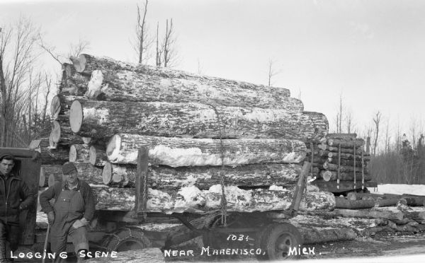 Two men stand next to a truck with a trailer full of snowy lumber, and more lumber in the background.