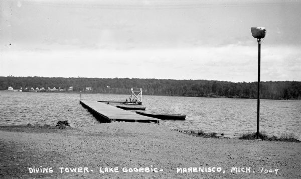 View looking at people on the dock and diving tower on Lake Gogebic.  Two boats are tied to the dock and buildings and trees are visible on the other side of the lake.