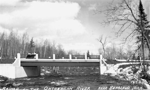 View from shoreline of a bridge over the Ontonagon River. There is snow on the ground with bare the trees in the background.