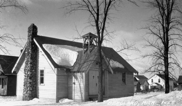 View of a one-story wooden church with a stone chimney and small bell tower over the entrance. Houses and trees are visible in the background, with snow on the ground.