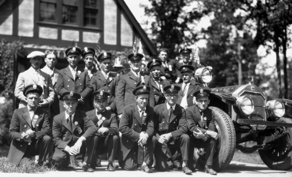 View of sixteen men members of the Montreal Fireman's Association, most wearing uniforms and hats, next to an early fire truck.