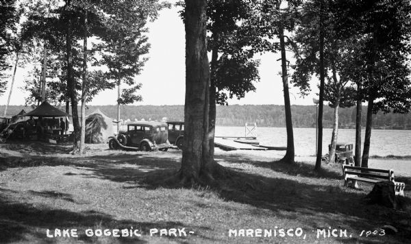 View of Lake Gogebic Park showing automobiles, trees, people around a picnic table, tents, a bench, dock and diving tower, as well as the lake and opposite shoreline visible in the distance.
