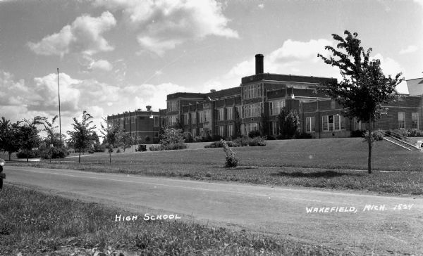 View of the Wakefield high school, a large brick building. Sections of the building are three stories high. There are some shrubs and small trees on the school lawn.