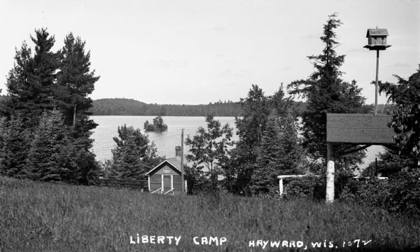 Small shack on shore of Little Spider Lake. Sign over door reads: "The Compleat Angler." There is a shelter made of birch tree trunks that has a birdhouse attached to the roof.