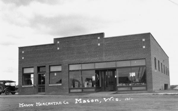 Exterior view of the brick front facade of the Mason Post Office and and Mason Mercantile Co. There is an automobile parked on the far left side of the building. There are two doorway entrances and on the front windows are the words “Post Office, Mason, Wis.”, “Mason Mercantile Co.” and “General Merchandise.”
