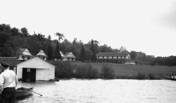 View of the White House Resort on Lake Gogebic. A man stands on a dock on the far left, while a wooden rowboat and boathouse are also visible in the foreground. There is a large building and four smaller cabins which are all a part of the resort on shore.