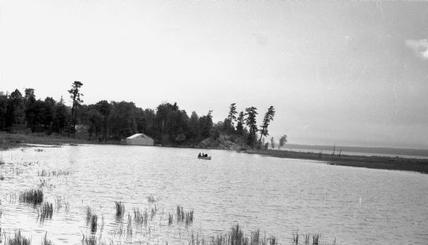 View across water of three people sitting in a wooden rowboat on Lake Gogebic near Marenisco. In the background is the shoreline, as well as a large building which may be a boathouse, and a dock.