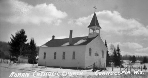 Exterior view of the Roman Catholic Church. It is a one-story wood, rectanglular building with a bell tower above the front entrance. There is snow on the ground and trees visible in the background.