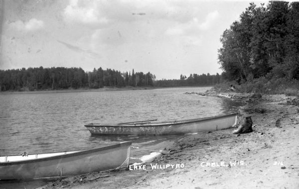 Two metal rowboats are pulled up on the shoreline of Lake Wilipyro.  The lake and trees on the opposite shore are visible in the background. A small animal, perhaps a dog, is on the shore at the far right facing away from the camera.