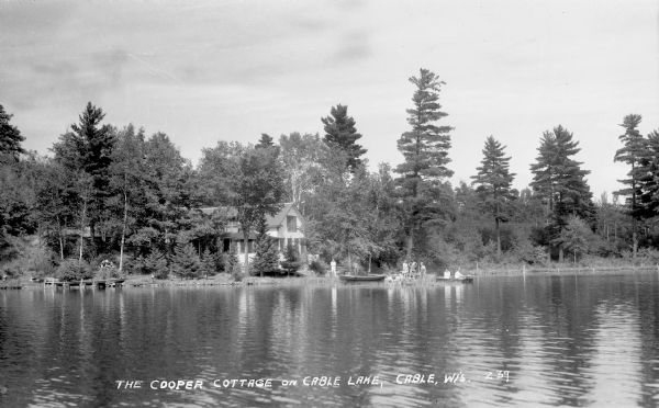 View across water of people standing on the shoreline and a dock. Two people are in a boat on the lake. Cooper Cottage is visible on land and another dock is on the far left.