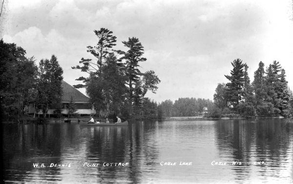 View across water of four people in a canoe on Cable Lake, with the W.A. Dennis Point Cottage and pine trees visible on the opposite shoreline.