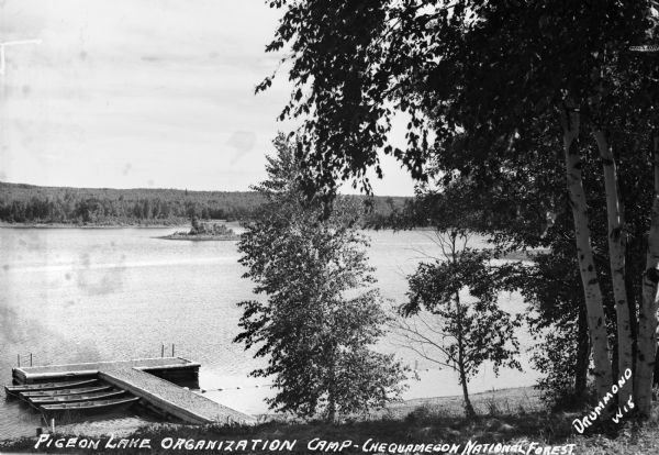 View down hill of Pigeon Lake with four boats at the dock. Trees surround the lake at Pigeon Lake Organization Camp in the Chequamegon National Forest.