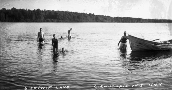 View of six boys and men swimming in Siskiwit Lake by a wooden boat. In the background is the shoreline and trees.