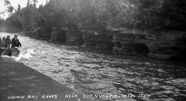 View of the Squaw Bay Caves, near Cornucopia, with people in a boat off to the far left. The caves were renamed Mawikwe Bay, and Mawikwe is Ojibwe for weeping woman.