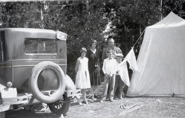 Outdoor family photograph of a mother, father, daughter and two sons.  They are standing in between a canvas tent and an automobile with a Saskatchewan license plate. In the background is the forest. The woman and girl are wearing dresses and hats, and the two boys have on dress shirts and bow ties. One boy wears a hat. The man is wearing suspenders and eyeglasses.