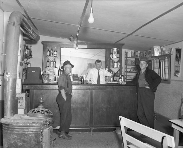 Interior view of a tavern with one man behind the bar and two men wearing hats standing at the bar holding bottles of beer. On the shelves behind the bar are bottles of liquor. In the foreground is a woodburning stove and a bench.