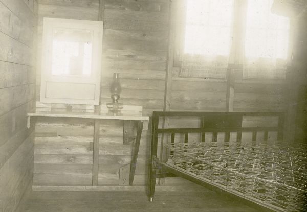 Interior view of a bedroom in a cottage at Nature Lovers Paradise on Jackson Lake, Bayfield County.In the bedroom a bed frame, box spring, end table and lamp are visible. Text with the photograph says, "Bedroom. Dressing table homemade with white oilcloth top. Plate glass mirror of finest quality. Shelves in medicine cabinet for toilet articles. 300 candle-power gas lamp for the main room; good quality oil lamps for night light in each bedroom. All windows have screens. Simmons steel beds. Finest double coil springs obtainable. Each bedroom holds two comfortably; and as there are two bedrooms each cottage will very COMFORTABLY hold four people --- 4 ladies; or 4 men; or two couples without children; or one couple with a maid or cook, or a family with children; if there are five in a family we will put an extra cot on the porch, but we will not rent a cottage for more than five people as it makes it too crowded. In case there are more than 5 to a party better rent two cottages and then invite enough to make the party number eight."<p>Located within the Chequamegon National Forest, Jackson Lake is a 142-acre drainage lake with a navigable outlet channel to Namakagon Lake – which is a 3,227 acre drainage lake at the headwaters of the Namakagon River in Bayfield County, Wisconsin.</p>