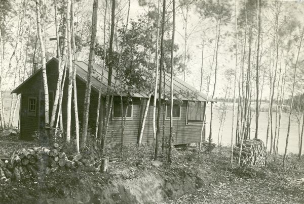 Exterior view of a cottage surrounded by birch trees at Nature Lovers Paradise on Jackson Lake near Grand View. Text with the photograph reads: "The BIRCHES – Each cottage is within fifty feet of the waters edge. (I mention this as I have seen LAKESHORE cottages that were over a half mile from the lakeshore). We furnish free firewood as shown in this photos; the wooden blocks are sawed right length; but the guest has to split them up; each cottage is furnish with an axe for this purpose. We have no garages for cars; but guest can drive his car up to the side of his cottage.  Rain don’t hurt a car that is closed up; and there is sufficient shade to protect the car from the sun that does the damage. Each cottage has a small but good 4 hole cast iron wood stove equipped with hot water reservoir. Water for washing is taken from the lake; water for drinking and cooking from a pump.  Besides regular cooking utensils each cottage has an enamel roaster, and cast iron pancake griddle and cast iron waffle iron (did you have these things at the last place you were at?)."<p>Located within the Chequamegon National Forest, Jackson Lake is a 142-acre drainage lake with a navigable outlet channel to Namakagon Lake – which is a 3,227 acre drainage lake at the headwaters of the Namakagon River in Bayfield County.