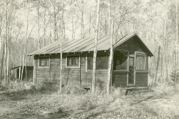 Exterior view of the Poplars Cottage surrounded by trees, part of Nature Lovers Paradise on Jackson Lake. Text above the photograph says, "The POPLARS --- Each cottage has a screened porch the full width of the front. Each cottage is equipped with one hammock. Toilets are of the outhouse type, kept clean. We would like to have electric lights, inside toilets, running water, etc. but we are just starting and cannot afford them yet as most of our money is invested in our land. However as much as we would like to have these things we do not consider them absolutely necessary. But good kitchen equipment to please the ladies and good beds to rest on; these we considered absolute necessities if our guests were going to have a good time while here; and we have gone the limit on these things and believe you will have a hard time to find better equipment anywhere, even in cottages renting for two and three times the price of ours."<p>Located within the Chequamegon National Forest, Jackson Lake is a 142-acre drainage lake with a navigable outlet channel to Namakagon Lake – which is a 3,227 acre drainage lake at the headwaters of the Namakagon River in Bayfield County.</p>