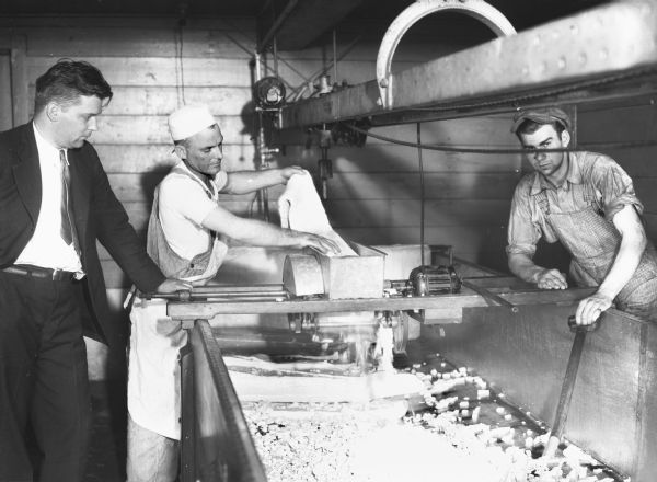 View of two men wearing work clothes, wearing overalls and hats, working at a machine, mostly likely making cheese curds. Another man wearing a suit and tie stands on the left watching the process.