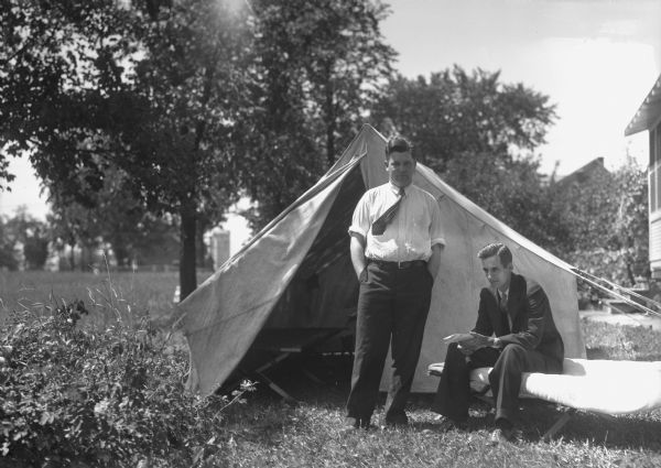 View of one man wearing a suit and tie sitting on a cot in front of a canvas tent. Another man wearing a dress shirt and tie is standing next to him in front of the tent doorway.