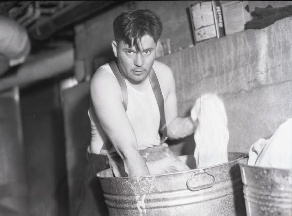Man looking at the camera while he is washing clothes in a washtub. He is wearing a sleeveless shirt and pants with suspenders while he does the laundry.