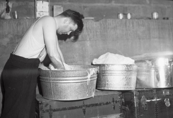 View of a man wearing a tank top and pants with suspenders washing clothing in three large metal washtubs, which are sitting on top of trunks.