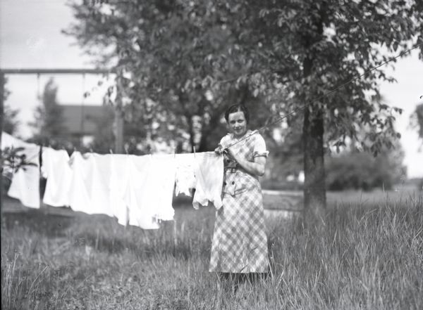 View of a woman wearing a plaid dress standing outdoors hanging up clean laundry, specifically light-colored shirts and towels, on the clothesline.