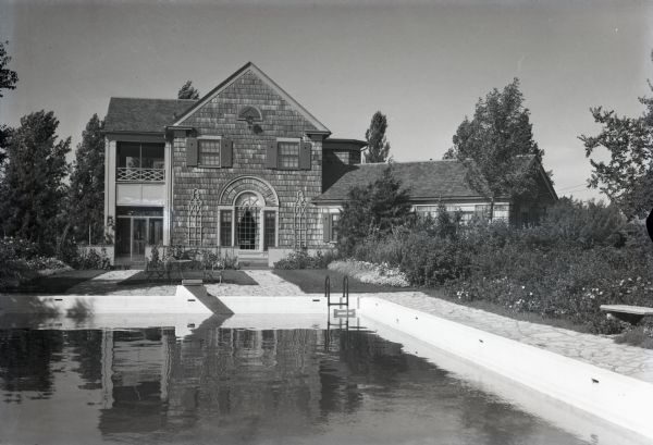 View of the two-story, wood shingle, lake shore home, Woods Manor, a part of Nebraska Row on Madeline Island. In the foreground is a swimming pool surrounded by gardens.