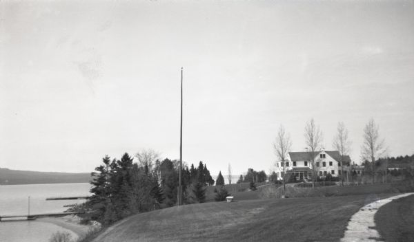 View of a house on Nebraska Row on Madeline Island. In the foreground is a flag pole, bench, path, lawn and trees. On the left are docks from the lake shore homes on Nebraska Row and Lake Superior. In the distance on the left is the Bayfield peninsula.