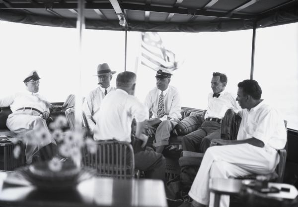 View of six men sitting on rattan furniture on a covered boat deck. Three men are wearing hats, and one man is smoking a cigar. Off the bow of the boat an American flag is flying.