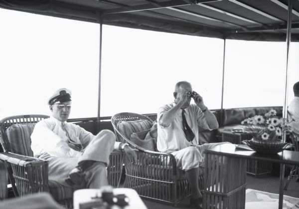 View of two men sitting in rattan chairs under a roof on the deck of a boat. They are both wearing dress shirts and ties. One man is wearing a hat, and the other man is holding binoculars up to his face.