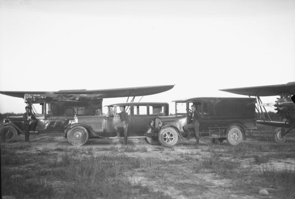 View of three young men, each standing in front of an automobile or truck. In the background and off to the right two airplanes are parked in the grass. The man on the left is holding a small dog. The truck on the right says "Goodwill Air Circus."