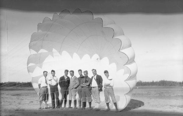 View of eight young men standing in a row facing the camera in front of a large open parachute. Some of the men are wearing tall boots, dress shirts, ties, and a few are wearing sweaters and jackets.