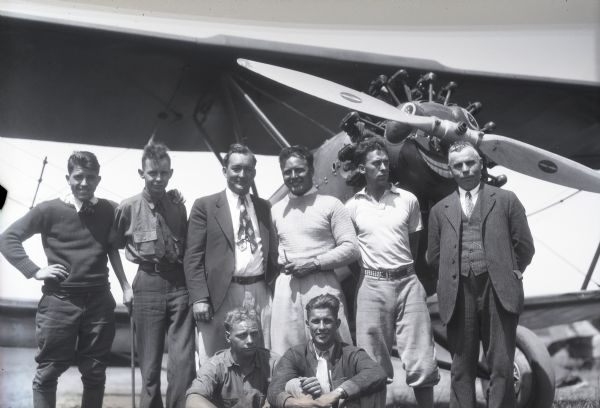 View of six men standing and two men sitting in front of an airplane.  The men are wearing a variety of clothing styles from work clothes to suits.