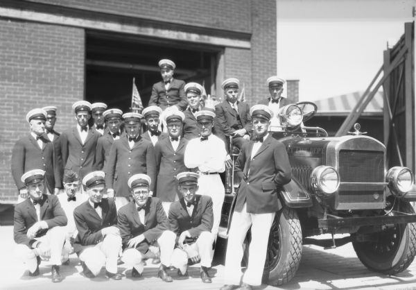 Group portrait of nineteen men of the Hurley Fire Department wearing hats and dress shirts. Many of the men are also wearing suit jackets. They are posed around a fire engine in front of the brick firehouse.