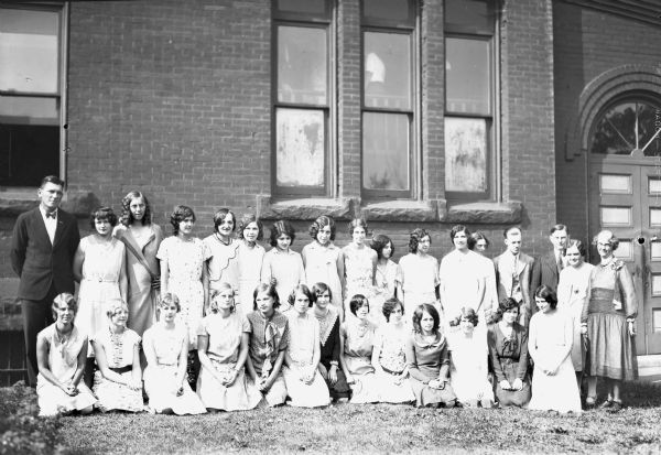 Group portrait of graduating students and teachers in front of a brick school building. There are twenty-seven women wearing dresses and three men wearing suits.