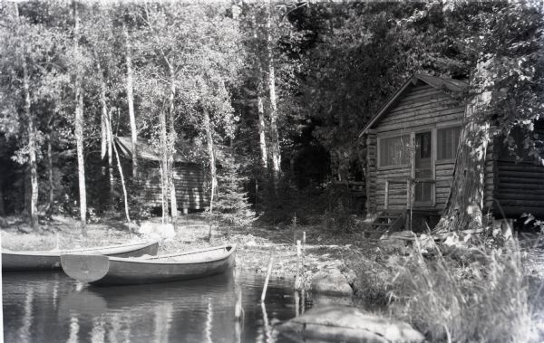 View from shoreline of two wooden boats tied up on the shoreline of  Les Crandall's Housekeeping Cottages on Lake Namakagon. On shore is a log cabin, a log outbuilding, trees and shrubs.