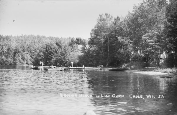View across water of people sitting in three boats next to a dock. People are standing on the dock, and one boat is pulled up on the shore. There are buildings barely visible through the trees in the background.