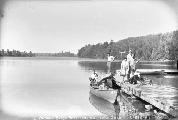 View of five people and a dog on the dock at the Virginia Beach boat landing. Two people are in a motor boat next to the dock. Another boat and dock is on the far right. Lake Owen and the opposite shoreline are in the background.