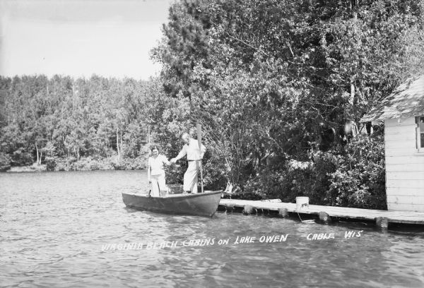 View of a man and woman getting into a wooden rowboat from a dock on Lake Owen at the Virginia Beach Cabins. Behind them a dog sits on the pier. On the far right is a boathouse. In the background is Lake Owen and trees on the opposite shoreline.