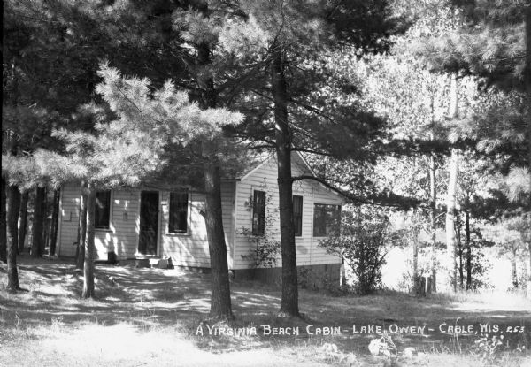 Exterior view of a small one-story wooden cabin with a porch, labeled as a Virginia Beach Cabin on Lake Owen. The cabin is surrounded by trees.