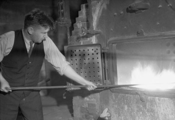 Interior view of a man wearing a vest and tie stoking a furnace.