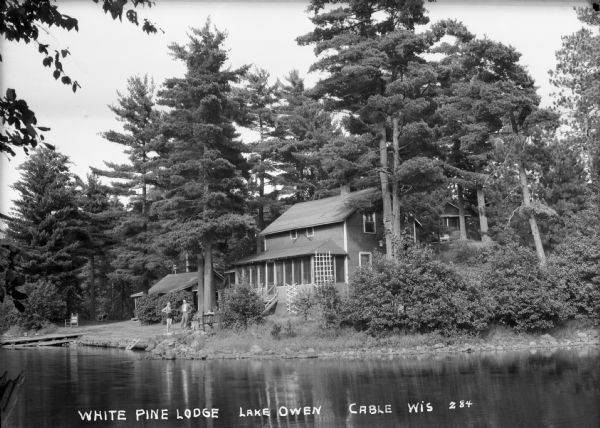 View across water of a man, woman, and two children on the shoreline of Lake Owen at the White Pine Lodge. There is a dock on the far left, and a two-story cabin with a front porch surrounded by tall trees. Other buildings are in the background.