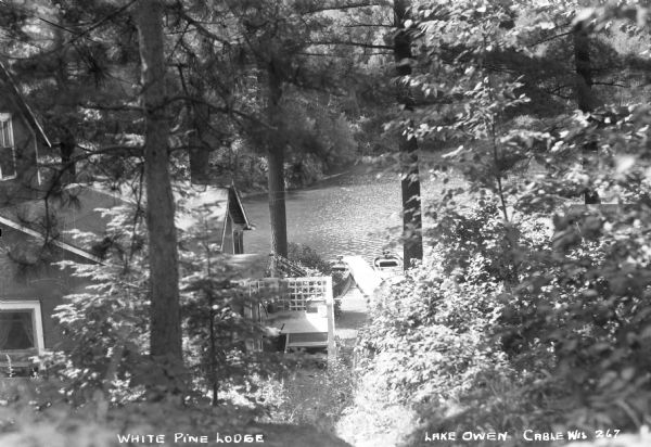 View looking down a hill at a cabins and a deck of the White Pine Lodge. On the shoreline of Lake Own is a dock with two boats.