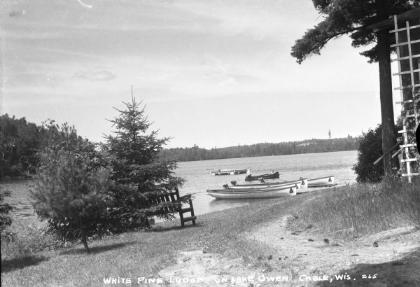 View of two people in a canoe on Lake Owen near White Pine Lodge. Just behind them  is a swimming platform. In the foreground is a dirt path in the grass, a bench, and trees.
